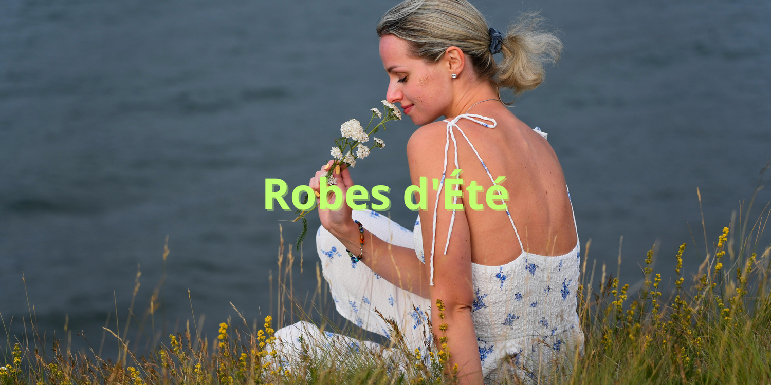 Femme en robe d'été blanche à fleurs bleues assise dans l'herbe au bord de l'eau, tenant une fleur.